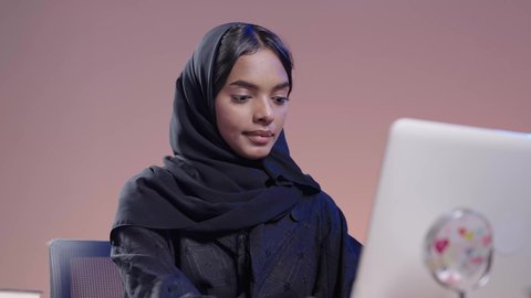 Integrating modern technologies into the educational field, dealing with online platforms in distance learning, portrait of a black female Saudi Arabian university student wearing an abaya and hijab sitting at a desk using a laptop, using modern technology in academic work