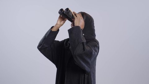 البحث وتوسيع مجال الرؤية باستخدام المنظار، إيماءات الاستغراب والمفاجأة، مفهوم الاستكشاف والمغامرة،طالبة جامعية سمراء عربيه سعوديه ترتدي العباءه والحجاب تقوم بالنظر من خلال المنظار ثنائي العينين ، خلفية بيضاء