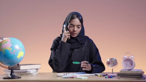 استخدام التكنولوجيا الحديثة في الأعمال الدراسيه، دمج التقنيات الحديثة بالمجال التعليمي،  بورتريه لطالبة جامعية سمراء عربيه سعوديه ترتدي العباءه والحجاب تجلس على مكتب بجانبها مجسم الكره الارضيه وتمسك الهاتف المحمول