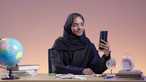 Integrating modern technologies into the educational field. Portrait of a black female Saudi university student wearing an abaya and hijab sitting at a desk next to a globe and holding a mobile phone. Using modern technology in academic work.