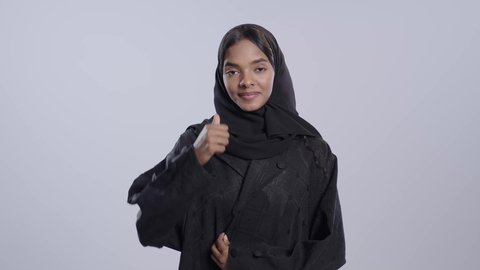 مفهوم القبول و الموافقة, تعابير الإعجاب و الرضى, طالبة جامعية سمراء عربيه سعوديه ترتدي العباءه والحجاب تنظر إلى الكاميرا و ترفع إصبع الإبهام للأعلى بإيماءات الجودة و الامتياز, خلفية ملونة