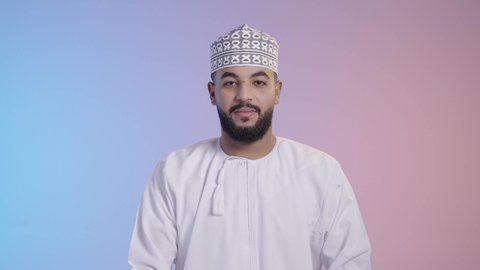 مفهوم الأناقة و الرجولة, الوقوف باسقامة و أيدي متقاطعة, الاهتمام بالمظهر الخارجي, بورتريه لرجل عربي خليجي عماني يرتدي الدشداشه والكمه ينظر إلى الكاميرا بإيماءات السعادة و السرور, خلفية ملونه