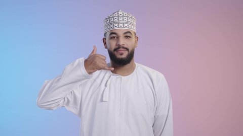 التواصل مع الأصدقاء ،مفهوم تلقي إتصال , إشارة تعني إتصل بي , بورتريه لرجل عربي عماني يرتدي الدشداشه والكمه, يقوم بإيماءات وجه ويدين تدل على إجراء مكالمه هاتفيه,  إنتظار مكالمه وإستخدام المايكروفون, خلفية ملونه