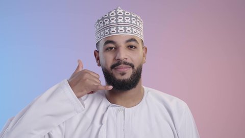 مفهوم تلقي إتصال , إشارة تعني إتصل بي , التواصل مع الأصدقاء ،بورتريه لرجل عربي عماني يرتدي الدشداشه والكمه, يقوم بإيماءات وجه ويدين تدل على إجراء مكالمه هاتفيه,  إنتظار مكالمه وإستخدام المايكروفون, خلفية ملونه