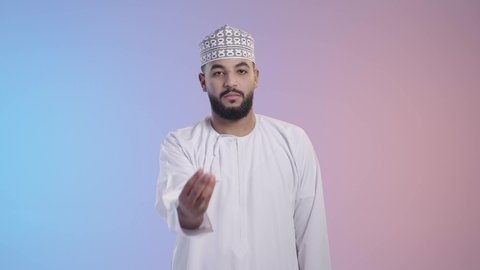 Giving time and waiting to do something, calm and asking to stop, portrait of an Arab Gulf Omani man wearing a dishdasha and a kemeh looking at the camera, gestures of patience, deliberation, and deliberation, colored background