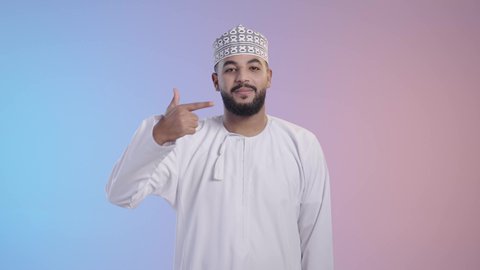 Oral hygiene and health care, avoiding cavities and gum disease, the concept of teeth whitening, beautification, and treatment. Portrait of a young Arab Gulf Omani man wearing a dishdasha and a sleeve pointing his index finger at his teeth, colored background.