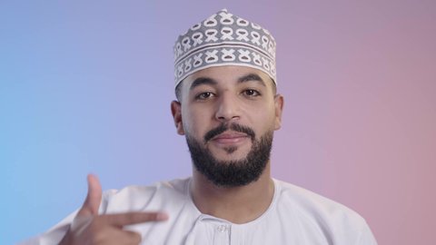 مفهوم تبييض وتجميل ومعالجة الأسنان, الاهتمام والعناية بنظافة وصحة الفم, تجنب التسوس وأمراض اللثة، بورتريه لشاب عربي خليجي عماني يرتدي الدشداشه والكمه يشير بإصبع السبابة إلى أسنانه, خلفية ملونه