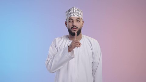 التوقف عن الكلام وخفض الصوت، طلب السكوت والصمت، الهدوء والسكون وعدم الإزعاج، رجل عربي خليجي عماني يرتدي الدشداشه والكمه يضع أصبع السبابة على فمه، حركة أصبع وإشارات، خلفية ملونه