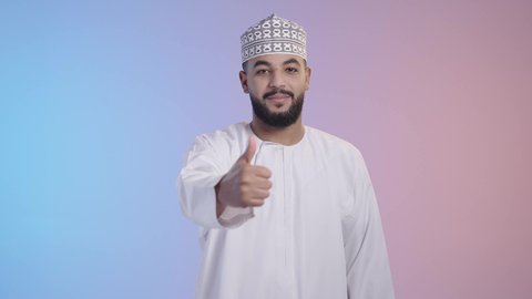 مفهوم القبول و الموافقة, تعابير الإعجاب و الرضى, شاب عربي خليجي عماني يرتدي الدشداشه والكمه ينظر إلى الكاميرا و يرفع إصبع الإبهام للأعلى بإيماءات الجودة و الامتياز, خلفية ملونة