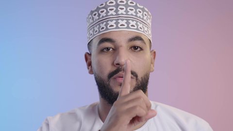 الهدوء والسكون وعدم الإزعاج، رجل عربي خليجي عماني يرتدي الدشداشه والكمه يضع أصبع السبابة على فمه، التوقف عن الكلام وخفض الصوت، طلب السكوت والصمت، حركة أصبع وإشارات، خلفية ملونه