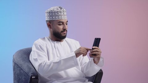 النظر إلى الهاتف المحمول بإيماءات الاندماج، تصفح مواقع وتطبيقات الانترنت، الدردشة والتحدث مع الأصدقاء، شاب عربي خليجي عماني يرتدي الدشداشه والكمه يمسك الجوال بيده، استخدام التقنيات والتكنولوجيا الحديثة بالحياة اليومية