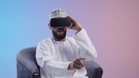 المتعة والتسلية بالمشاهدة عبر نظارة الواقع الافتراضي, رجل عربي خليجي عماني يرتدي الدشداشه والكمه يجلس على كرسي يضع نظارات ثلاثية الأبعاد يقوم برفع كف يده للاعلى بتعابير الاندماج , ايماءات التوقف عن فعل امر ما
