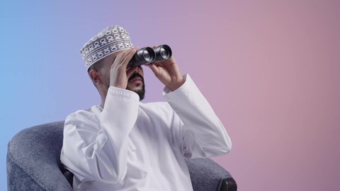 Exploring and expanding the field of vision using binoculars, gestures of wonder and surprise, the concept of exploration and adventure, a young Arab Gulf Omani man wearing a dishdasha and a cloak looking through binoculars, colored background