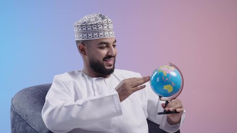Focus gestures and the concept of searching and exploring the world. Pointing to a specific location on a map and looking at a continent. Portrait of an Omani Gulf Arab man wearing a dishdasha and a kimmah holding a globe. Colorful background.