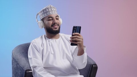 استخدام التقنيات الحديثه ، الاستماع للأغاني والمواهب الغنائية عبر الجوال ،شاب عربي خليجي عماني يرتدي الدشداشه والكمه يضع سماعات الرأس ويمسك بيده الهاتف المحمول بإيماءات الفرح والسرور، مفهوم الإصغاء باستخدام الهيدفون،التفاعل والاندماج مع الأنغام الموسيقية,  خلفية ملونة