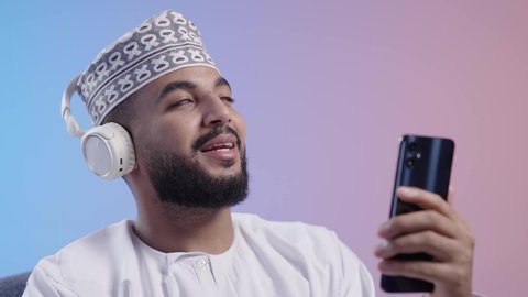 الاستماع للأغاني والمواهب الغنائية عبر الجوال ،شاب عربي خليجي عماني يرتدي الدشداشه والكمه يضع سماعات الرأس ويمسك بيده الهاتف المحمول بإيماءات الفرح والسرور، مفهوم الإصغاء باستخدام الهيدفون،التفاعل والاندماج مع الأنغام الموسيقية,  خلفية ملونة