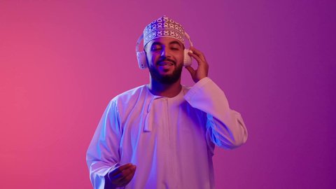 شاب عربي خليجي عماني يرتدي الدشداشه والكمه يضع سماعات الرأس بإيماءات الفرح والسرور، مفهوم الإصغاء باستخدام الهيدفون، الاستماع للأغاني والمواهب الغنائية،التفاعل والاندماج مع الأنغام الموسيقية,  خلفية ملونة