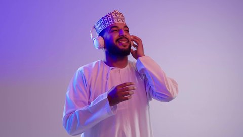 مفهوم الإصغاء باستخدام الهيدفون، الاستماع للأغاني والمواهب الغنائية،التفاعل والاندماج مع الأنغام الموسيقية,  شاب عربي خليجي عماني يرتدي الدشداشه والكمه يضع سماعات الرأس بإيماءات الفرح والسرور، خلفية ملونة