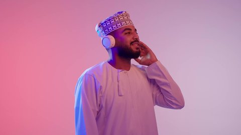 الاستماع للأغاني والمواهب الغنائية، مفهوم الإصغاء باستخدام الهيدفون، التفاعل والاندماج مع الأنغام الموسيقية,  شاب عربي خليجي عماني يرتدي الدشداشه والكمه يضع سماعات الرأس بإيماءات الفرح والسرور، خلفية ملونة