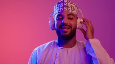 التفاعل والاندماج مع الأنغام الموسيقية, الاستماع للأغاني والمواهب الغنائية، مفهوم الإصغاء باستخدام الهيدفون، شاب عربي خليجي عماني يرتدي الدشداشه والكمه يضع سماعات الرأس بإيماءات الفرح والسرور، خلفية ملونة