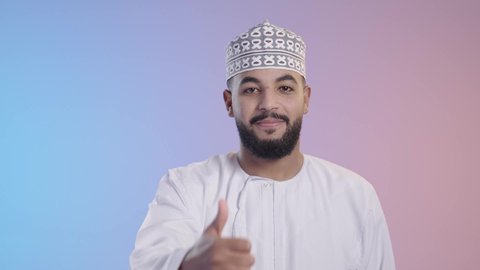 The concept of acceptance and approval, expressions of admiration and satisfaction, a young Arab Gulf Omani man wearing a dishdasha and a kemeh looking at the camera and giving a thumbs-up gesture of quality and excellence, on a colorful background.