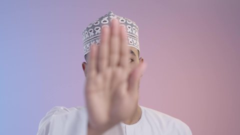Request to refrain from approaching, to protect oneself from external harm. Photograph of a young Arab Gulf Omani man wearing a dishdasha and a keema, looking at the camera and raising his hand with an expression of disapproval, indicating disapproval. Colored background.