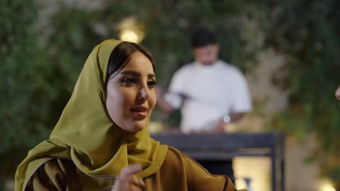 قضاء وقت ممتع ومسلي مع مجموعة من الأصدقاء،  اجتماع الأهل والأصدقاء في الهواء الطلق، إمرأة عربية خليجية سعوديه ترتدي الزي التقليدي تمسك كأس به مشروب بارد تقوم بالتحدث مع الأصدقاء بإيماءات الاندماج، التنزه والتسلية والترفيه في شاليه سياحية في السعوديه