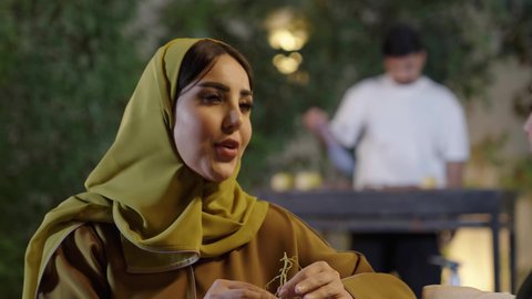 اجتماع الأهل والأصدقاء في الهواء الطلق، قضاء وقت ممتع ومسلي مع مجموعة من الأصدقاء، إمرأة عربية خليجية سعوديه ترتدي الزي التقليدي تمسك كأس به مشروب بارد تقوم بالتحدث مع الأصدقاء بإيماءات الاندماج، التنزه والتسلية والترفيه في شاليه سياحية في السعوديه