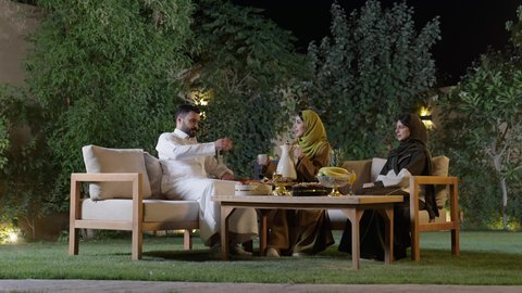اجتماع الأهل والأصدقاء في الهواء الطلق، أصدقاء عربيون خليجيون سعوديون بالزي التقليدي يشربون القهوة العربية ويتبادلون الأحاديث والقصص بإيماءات الاندماج، التنزه والتسلية في شاليه سياحية في وقت العطلة ، تجسيد مفهوم حسن الضيافة والكرم العربي،دلة القهوة العربية تراث قديم، قضاء وقت ممتع ومسلي مع مجموعة من الأصدقاء