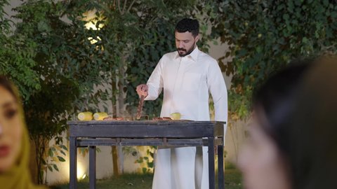 شاب سعودي يقوم بعملية الشواء تقليب اللحم على الموقد ، إمرأة عربية خليجية سعوديه ترتدي الزي التقليدي تمسك كأس به مشروب بارد تقوم بالتحدث مع الأصدقاء بإيماءات الاندماج، التنزه والتسلية والترفيه في شاليه سياحية في السعوديه، اجتماع الأهل والأصدقاء في الهواء الطلق، قضاء وقت ممتع ومسلي مع مجموعة من الأصدقاء