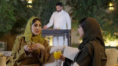 التنزه والتسلية والترفيه في شاليه سياحية في السعوديه، اجتماع الأهل والأصدقاء في الهواء الطلق، قضاء وقت ممتع ومسلي مع مجموعة من الأصدقاء، إمرأة عربية خليجية سعوديه ترتدي الزي التقليدي تمسك كأس به مشروب بارد تقوم بالتحدث مع الأصدقاء بإيماءات الاندماج