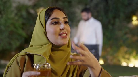 قضاء وقت ممتع ومسلي مع مجموعة من الأصدقاء، اجتماع الأهل والأصدقاء في الهواء الطلق، تصوير مقرب لإمرأة عربية خليجية سعوديه ترتدي الزي التقليدي تمسك كأس به مشروب بارد تقوم بالتحدث مع الأصدقاء بإيماءات الاندماج، التنزه والتسلية والترفيه في شاليه سياحية في السعوديه