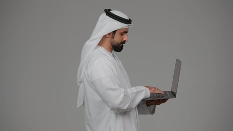 ادارة شؤون الاعمال وتنظيمها، العمل عن بعد، تصفح مواقع التواصل الإجتماعي ،استخدام الأجهزة التقنية الحديثة والمتطورة، بورتريه من الجانب لرجل عربي خليجي اماراتي يرتدي الكندورة والغترة البيضاء يستخدم الحاسوب المحمول، اجراء مكالمات الفيديو، خلفية رمادية