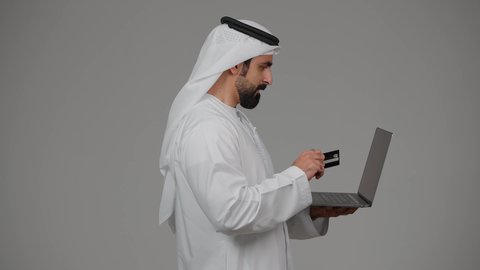 مفهوم الدفع والتسديد عن بعد عبر تطبيقات اللابتوب، استخدام البطاقة البنكية الالكترونية، تطور التكنولوجيا والأجهزة التقنية، بورتريه من الجانب لرجل عربي اماراتي خليجي يرتدي الكندورة والغترة يعمل على جهاز الحاسوب المحمول، خلفية رمادية