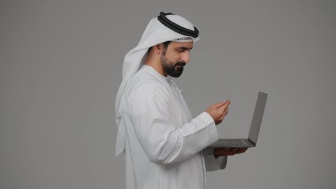 مفهوم الدفع والتسديد عن بعد عبر تطبيقات اللابتوب، استخدام البطاقة البنكية الالكترونية، تطور التكنولوجيا والأجهزة التقنية، بورتريه من الجانب لرجل عربي اماراتي خليجي يرتدي الكندورة والغترة يعمل على جهاز الحاسوب المحمول، خلفية رمادية