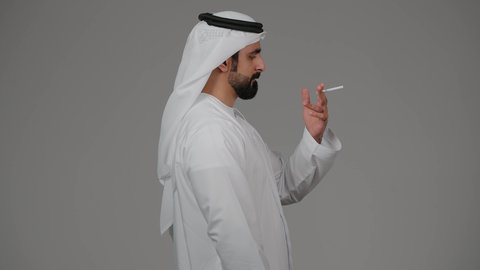 الادمان على التبغ و السجائر، عادات سلوكية ضارة، مفهوم مكافحة التدخين, تعاطي مواد سامة، بورتريه من الجانب لرجل عربي اماراتي خليجي يرتدي الكندورة والغترة يقوم بتدخين السجائر، خلفيه رمادية