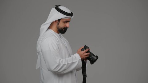 احتراف التصوير الفوتوغرافي باستخدام الكاميرات الرقمية, ايماءات وتعابير السرور والسعادة،بورتريه من الجانب لشاب عربي خليجي اماراتي مبتسم يرتدي الكندورة والغترة يقوم بالتقاط صور تذكارية عبر كاميرا فوتوغرافية, ممارسة هواية التصوير, خلفية رمادية
