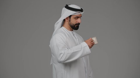 تأثير الكافيين على نشاط الدماغ والذاكرة، الاستمتاع بتناول المشروبات الساخنة والأعشاب الشتوية الدافئة، بورتريه من الجانب لرجل عربي خليجي اماراتي يرتدي الكندورة والغترة يقوم بشرب فنجان من القهوة، خلفية رمادية