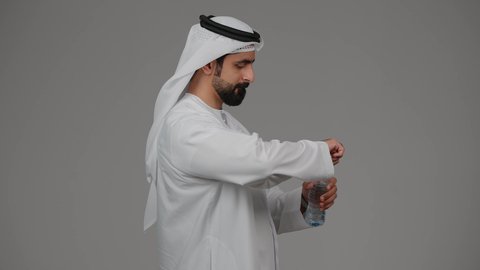 انتظار موعد محدد، مفهوم اهميه الوقت والالتزام بالمواعيد، بورتريه من الجانب لرجل عربي خليجي اماراتي يرتدي الدكندورة والغترة ينظر الى ساعته الشخصيه بايماءات اهميه الالتزام بالوقت وإنجاز المهامات والاعمال بدقه، خلفيه رمادية