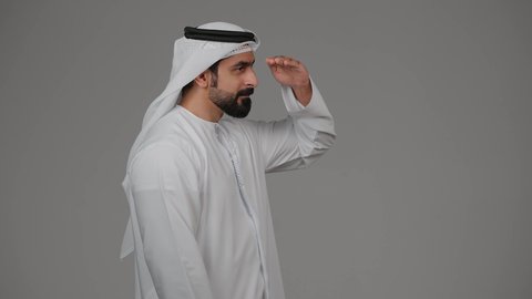 مفهوم التطلع للمستقبل, النظر لمكان ما, تعابير محاولة رؤية الأشياء البعيدة,بورتريه من الجانب لرجل عربي خليجي إماراتي يرتدي الكندورة الغترة البيضاء يرفع يده إلى رأسه بإيماءات البحث عن شيء محدد, خلفية رمادية
