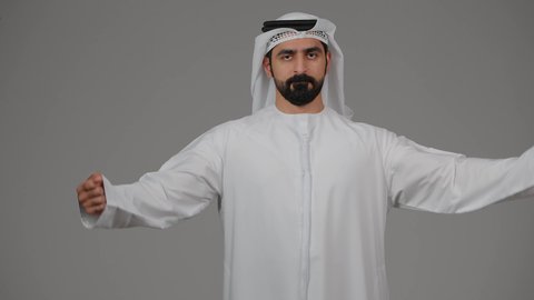 اللياقة البدنية والجسم الصحي المتين، التمتع بالبنية الجسدية القوية، النظر إلى الكاميرا بإيماءات الثقة بالنفس، بورتريه لرجل عربي خليجي اماراتي يرتدي الكندورة والغترة البيضاء يقوم بعرض عضلات يديه، إيماءات تدل على القوة العضلية،  خلفية رمادية