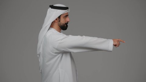 تحديد موقع شيء معين، إشارات وحركات يدين تدل على هذا المكان (هنا)، بورتريه من الجانب لرجل عربي خليجي اماراتي يرتدي الكندورة والغترة البيضاء يشير باصبع السبابة إلى شيء ما، مفهوم الأناقة والاهتمام بالمظهر الخارجي، خلفية رمادية