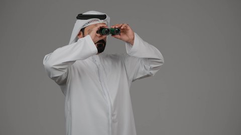 حب المغامرة والاستكشاف، رؤية الاشياء البعيدة عبر التلسكوب، نظرة ورؤية مستقبلية، بورتريه لرجل عربي خليجي إماراتي يرتدي الكندورة والغترة ينظر من خلال المنظار ثنائي العينين، خلفية رمادية