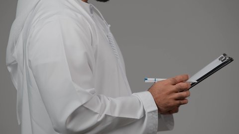 تنظيم وإدارة الأعمال ، كتابة خطة العمل، تدوين وتسجيل الملاحظات والمعلومات على الورق, بورتريه من الجانب لشاب عربي خليجي اماراتي يرتدي الكندورة والغترة يقوم بكتابة المهام والأهداف اليومية, خلفية رمادية
