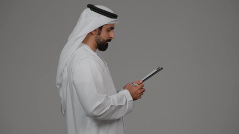 تنظيم وإدارة الأعمال ، كتابة خطة العمل، تدوين وتسجيل الملاحظات والمعلومات على الورق, بورتريه من الجانب لشاب عربي خليجي اماراتي يرتدي الكندورة والغترة يقوم بكتابة المهام والأهداف اليومية, خلفية رمادية