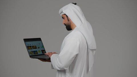 Business management and organization, remote work, browsing social media, using modern and advanced technical devices, side portrait of an Emirati Gulf Arab man wearing a kandura and ghutra using a laptop, making video calls, gray background