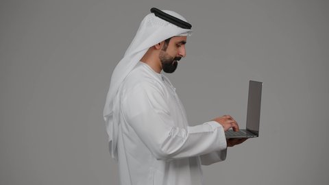 ادارة شؤون الاعمال وتنظيمها، العمل عن بعد، تصفح مواقع التواصل الإجتماعي ،استخدام الأجهزة التقنية الحديثة والمتطورة، بورتريه من الجانب لرجل عربي خليجي اماراتي يرتدي الكندورة والغترة يستخدم الحاسوب المحمول، اجراء مكالمات الفيديو، خلفية رمادية