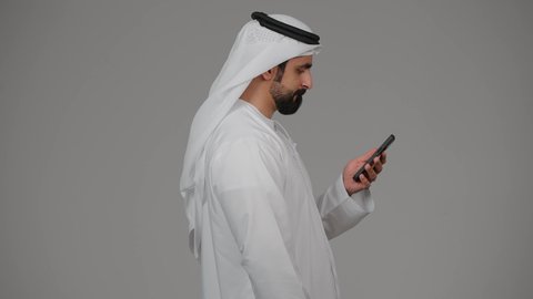 Integrating modern technology into daily life, communicating with family and friends, making a video call, taking selfies with a mobile phone, documenting personal memories, side portrait of an Emirati Gulf Arab man wearing a kandura holding a mobile phone, gray background.