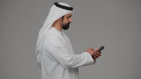 دمج التقنيات والتكنولوجيا الحديثة بالحياة اليومية،  التواصل مع الاهل والاصدقاء، اجراء مكالمة فيديو ،التقاط صور السيلفي باستخدام الهاتف المحمول، توثيق اللقطات الشخصية التذكارية، بورتريه من الجانب لرجل عربي خليجي اماراتي يرتدي الكندورة يمسك الجوال ، خلفية رمادية.