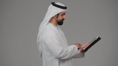عقد اجتماع عمل عن بعد عبر الجهاز اللوحي، التواصل مع الاهل والاصدقاء، دمج التقنيات والتكنولوجيا الحديثة بالحياة اليومية، بورتريه من الجانب لرجل عربي خليجي اماراتي يرتدي الكندورة و الغترة يقوم بإجراء مكالمة فيديو عبر التابليت المحمول، خلفية رمادية.